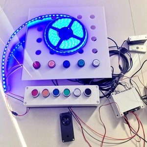 Puede incluir: Un panel de control blanco con seis botones de colores y una caja blanca con cables. Una tira LED azul está enrollada alrededor de un carrete y un altavoz está sobre la mesa.