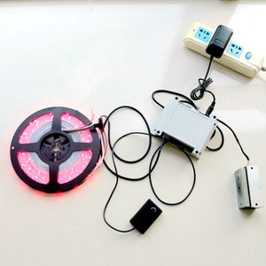 May include: A white power adapter plugged into a power strip with a black power cord connected to a white box with a black wire connected to a roll of pink LED lights. A black remote control is also included.