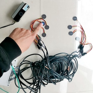 May include: A close-up of a black and white touch sensor pad with a blue light indicating activation. The pad is connected to a black box with wires and a tangle of black cables.