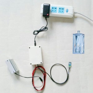 Op de afbeelding: Een witte stroomadapter die is aangesloten op een witte stekkerdoos met drie stopcontacten. Een witte doos met draden die eraan zijn bevestigd, bevindt zich aan de linkerkant van de afbeelding. Een zwarte draad verbindt de doos met een klein blauwgroen apparaat. Een witte lantaarn met een transparant glazen paneel bevindt zich aan de rechterkant van de afbeelding.