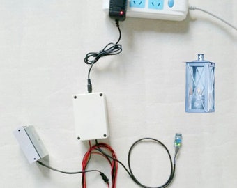 Puede incluir: Un adaptador de corriente blanco enchufado a una regleta blanca con tres enchufes. Una caja blanca con cables conectados a ella se encuentra en el lado izquierdo de la imagen. Un cable negro conecta la caja a un pequeño dispositivo azul y verde. Una linterna blanca con un panel de vidrio transparente se encuentra en el lado derecho de la imagen.