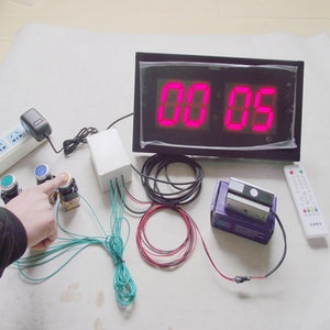 Puede incluir: Un temporizador de cuenta atrás digital rojo con marco negro, una caja de control blanca, un mando a distancia y tres botones verdes y azules. El temporizador muestra "00:00".
