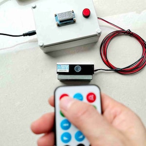 Pode incluir: Uma caixa branca com um botão vermelho e um visor digital de temporizador, uma caixa preta com um rótulo, um controle remoto com botões vermelhos, azuis e brancos e fios vermelhos e pretos.