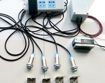 Puede incluir: Un conjunto de cuatro sensores de proximidad negros y plateados con puntas azules, cuatro soportes de montaje metálicos, un adaptador de corriente negro, una regleta de enchufes blanca con dos tomas de corriente, una caja eléctrica gris y un interruptor magnético negro.