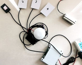 Puede incluir: Un dispositivo blanco, redondo, inalámbrico con cables negros conectados a él. Hay cuatro tarjetas blancas con los números 2, 3 y 4 escritos en ellas. Hay una caja negra con un botón blanco y una caja blanca con una placa de circuito verde.