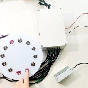 May include: A white circular button pad with 12 silver buttons, one of which is being pressed by a finger. The button pad is connected to a white box with wires.