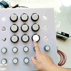 May include: A white panel with a grid of 15 round, black-rimmed buttons. One button is lit up, and a finger is pointing at it.