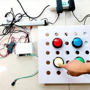 Puede incluir: Una tabla blanca con una cuadrícula de agujeros y cuatro botones redondos. Los botones son rojo, azul, amarillo y verde. Una mano está presionando el botón amarillo.