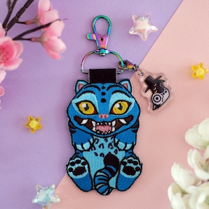 May include: A blue embroidered keychain featuring a cartoon cat with yellow eyes and an open mouth. The keychain has a rainbow-colored clasp and a small clear charm. The background is a mix of purple and pink with floral and star decorations.