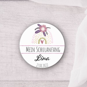 Könnte beinhalten: Ein weißer runder Aufkleber mit einem rosa, violetten und goldenen Blumendesign. Der Text "Mein Schulanfang" ist in Schwarz auf einem rosa Banner geschrieben. Der Name "Lina" ist in schwarzer Kursivschrift unter dem Banner geschrieben. Das Datum "27.08.2022" ist in Schwarz unter dem Namen geschrieben.