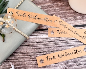 Puede incluir: Un regalo envuelto en papel azul claro, adornado con una ramita verde, una bola de cristal transparente con estrellas doradas y una cuerda dorada. La etiqueta del regalo dice "Frohe Weihnachten". Velas y otras decoraciones en el fondo.