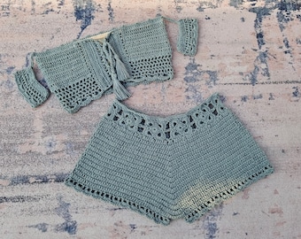 May include: A crocheted two-piece set in a sage green color. The top is a halter style with a tie closure and a tassel. The shorts are high-waisted and have a scalloped edge.