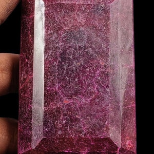 May include: A large, deep red, faceted gemstone with a rectangular shape. The gemstone has a slightly cloudy appearance with visible internal lines.