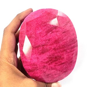 Puede incluir: Una gran gema de rubí ovalada y facetada. El rubí es de un tono rojo rosado vibrante con texturas internas visibles y reflejos de luz. La gema se sostiene en una mano sobre un fondo blanco.