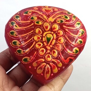 May include: A heart-shaped box with a glossy red finish, featuring a hand-painted decorative pattern in shades of yellow, orange, and green. The intricate design radiates from the center, covering the entire top surface of the small container.