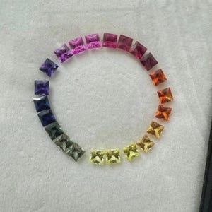 Può includere: Una collezione di gemme a taglio quadrato disposte in un motivo circolare, che mostra uno spettro di colori dal viola al giallo. Le gemme sono esposte su una superficie chiara.