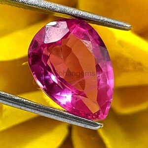 Puede incluir: Una piedra preciosa rosa en forma de pera con facetas, sostenida por unas pinzas sobre un fondo amarillo.