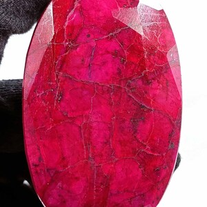 Könnte beinhalten: Ein großer, ovaler, tief roter Rubin-Edelstein mit facettiertem Schliff. Der Rubin hat ein natürliches, leicht trübes Aussehen mit sichtbaren Einschlüssen.