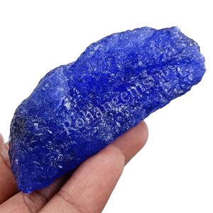 Può includere: Una gemma grezza, blu intenso, con una superficie ruvida e strutturata. La pietra è tenuta in mano, mostrando la sua forma e il suo colore naturali. Il testo "Rohangems" è visibile.