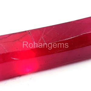 May include: A deep red, faceted, rough ruby gemstone. The ruby is a rectangular shape with a smooth, polished surface. The ruby is approximately 2 inches long.