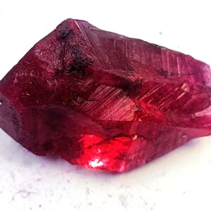 May include: A raw, uncut ruby gemstone. The ruby is a deep, saturated red color with visible crystalline structures and a polished, reflective surface. The gemstone is set against a white background.