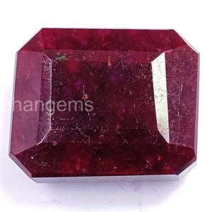 May include: A faceted, rectangular ruby gemstone with a deep red hue. The gem has a polished surface with visible internal inclusions and a slight sparkle. The word "hangems" is visible on the surface of the gem.