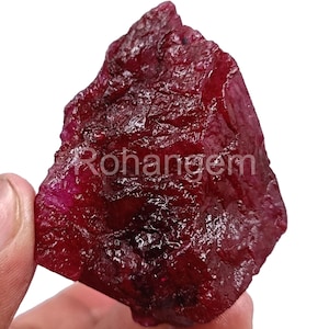May include: A rough-cut, deep red ruby gemstone with a textured surface. The ruby is held in a hand, showcasing its natural, irregular shape. The text "Rohangem" is visible on the surface of the gemstone.