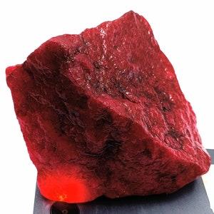 Può includere: Una grande gemma rossa grezza con un colore profondo e ricco. La gemma è illuminata dal basso, proiettando un bagliore caldo.
