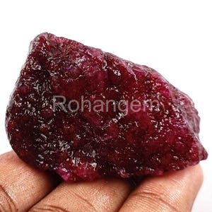 May include: A raw, uncut ruby gemstone in a deep red hue. The stone's surface has a textured appearance. The word "Rohangem" is visible on the surface. The gemstone is held in a hand.