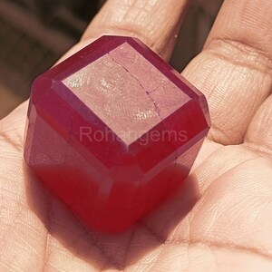 Op de afbeelding: Een gefacetteerde, dieprode edelsteen met een rechthoekige vorm. De edelsteen wordt in een hand gehouden, waarbij de gepolijste oppervlakken en interne helderheid worden getoond. De tekst "Rohangems" is zichtbaar op de edelsteen. Deze edelsteen is voor sieraden.