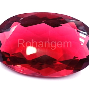 May include: A faceted, oval-shaped gemstone with a deep red hue. The gem has a polished surface that reflects light, creating a sparkling effect. The word "Rohangem" is printed on the surface of the gem.