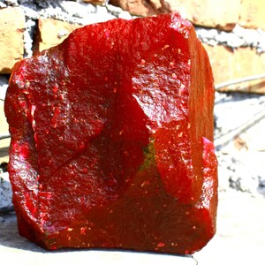 Puede incluir: Una piedra preciosa roja grande, áspera, con una forma ligeramente irregular. La piedra preciosa tiene un color rojo intenso y rico y un acabado brillante.