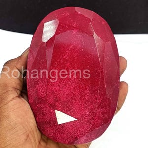May include: A large, faceted, oval-shaped ruby gemstone. The ruby is a deep red color with a polished surface. The text "Rohangems" is visible on the left side of the gemstone. A white triangle shape is visible on the bottom of the gemstone.
