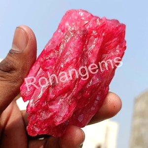 May include: A large, rough, red crystal with a layered, textured surface. The crystal is held in a hand, and the background is a blue sky.