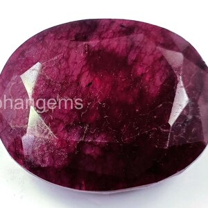 Puede incluir: Gema de rubí de corte ovalado con un tono rojo intenso. La gema tiene múltiples facetas y una superficie pulida, que refleja la luz. El texto "ohangems" es visible en la superficie. Adecuado para la creación de joyas.