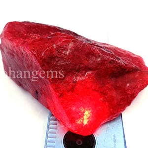 May include: A large, rough-cut ruby gemstone with a deep red color. The gemstone has a textured surface and is positioned on a light blue surface. The word "hangems" is visible on the gemstone.