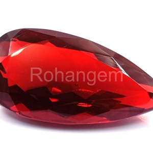 May include: A faceted, teardrop-shaped gemstone in a deep red hue. The gem has a polished surface with multiple facets, reflecting light. The word "Rohangem" is printed on the surface. The gemstone is set against a white background.