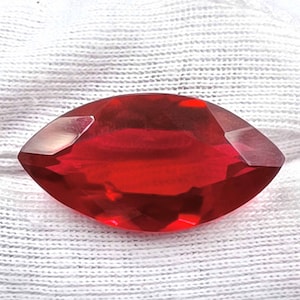 Op de afbeelding: Een diep rode, gefacetteerde edelsteen met een marquise-vorm. De edelsteen ligt op een witte stoffen achtergrond.