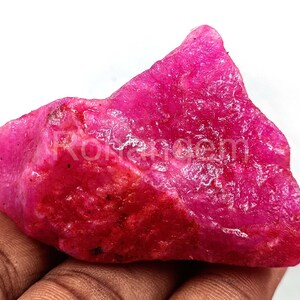 Puede incluir: Una gema de rubí en bruto, sin cortar, con un vibrante tono rojo rosado. La piedra natural tiene una superficie texturizada y una forma irregular, mostrando su estado bruto y sin pulir. La imagen destaca el color y la estructura cristalina de la gema.