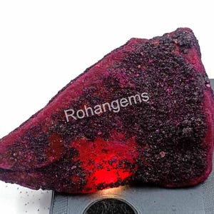Könnte beinhalten: Ein großer, roher, roter Rubin-Edelstein mit schwarzen Einschlüssen. Der Rubin wird von unten beleuchtet, wodurch seine rote Farbe hervorgehoben wird. Der Text "Rohangems" ist auf dem Rubin sichtbar.