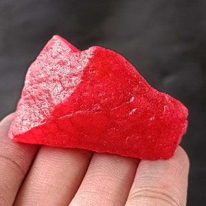 May include: A large, bright red, irregularly shaped crystal. The crystal has a smooth, polished surface and is held in a hand.