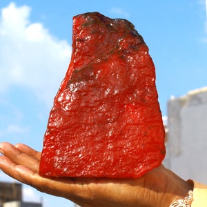 May include: A large, irregularly shaped, red gemstone with a smooth, polished surface. The gemstone is being held in a person's hand.