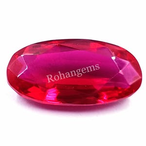 Puede incluir: Una gema ovalada de color rojo vibrante con un corte facetado. La gema está colocada sobre un fondo blanco. Rohangems está impreso en la gema.