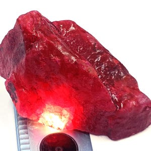 May include: A raw, uncut ruby gemstone with a deep red hue. The rough stone has a textured surface and irregular shape, with areas of lighter and darker coloration. The ruby is positioned on a silver measuring device.