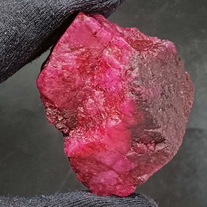 May include: A large, rough, red ruby gemstone with a dark red to black hue on one side. The ruby is a natural, unpolished stone.