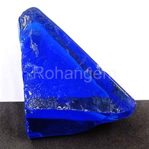 May include: A large, irregularly shaped piece of deep blue glass. The translucent material has a rough, textured surface with sharp edges and a glossy sheen. The word "Rohangem" is visible on the surface. The object is set against a white and black background.