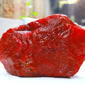 Può includere: Una grande gemma rossa grezza con una superficie lucida. La pietra ha una forma irregolare e sembra essere un esemplare naturale non tagliato. Il colore è un rosso intenso e vibrante.