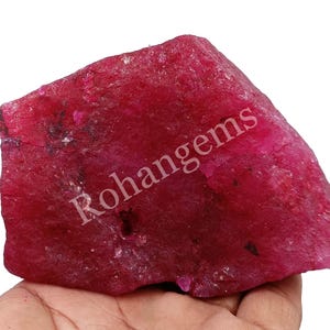 Puede incluir: Una gema de rubí en bruto, sin cortar, de color rojo intenso. La superficie tiene un aspecto texturizado, y la palabra "Rohangems" está impresa en la piedra. La gema se sostiene en una mano.