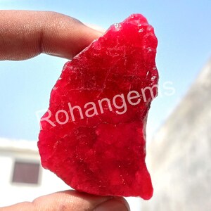 May include: A large, rough, red ruby gemstone. The ruby is translucent and has a deep red colour. The ruby is being held by a hand.