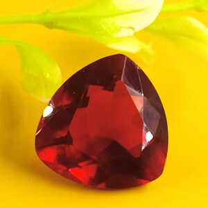 May include: A deep red, faceted gemstone with a triangular shape. The gemstone is set against a bright yellow background.